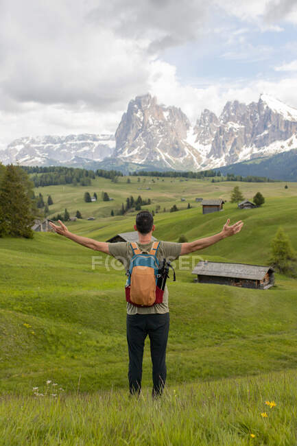 Vista posterior de un viajero anónimo con una mochila de pie con los brazos extendidos y disfrutando de la libertad en Seiser Alm en las montañas Dolomite - foto de stock