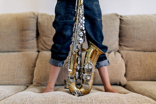 Recortado anónima niño descalzo reflexivo en sombrero con saxofón de pie en el sofá contra la ventana en casa - foto de stock