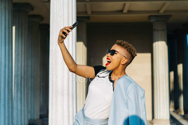 Mulher afro-americana adulta elegante com corte de cabelo moderno e jaqueta tirando selfie no celular contra a parede de azulejos com sombra na luz solar — Fotografia de Stock