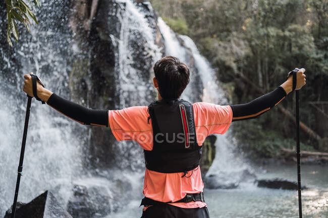 Vista posteriore dell'escursionista maschio con bastoni da trekking in mani distese in piedi con gli occhi chiusi vicino alla cascata nel bosco e godendo della libertà — Foto stock