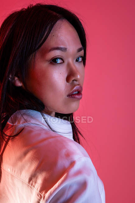 Vista lateral de modelo feminino étnico confiante em roupas da moda olhando para a câmera em estúdio no fundo rosa — Fotografia de Stock