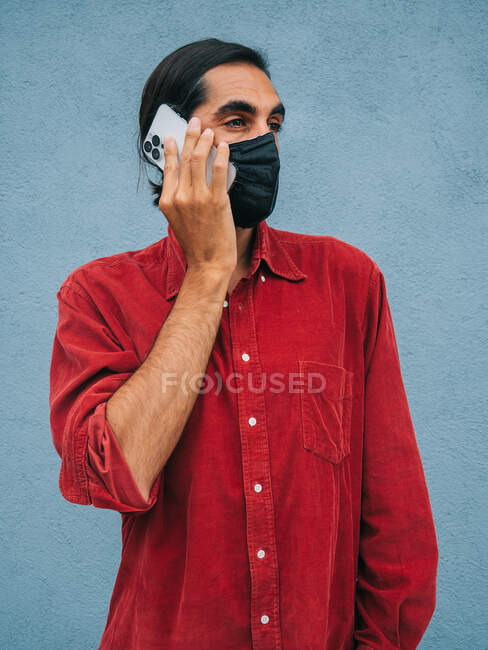 Спокійний етнічний чоловік у захисній масці, який розмовляє по мобільному телефону, стоячи проти синьої стіни в місті під час коронавірусу. — стокове фото