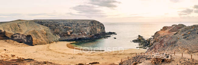 Amplia vista angular de la tranquila laguna con agua de mar clara cerca de la playa de arena y acantilados contra el cielo nublado del amanecer en Fuerteventura, España - foto de stock