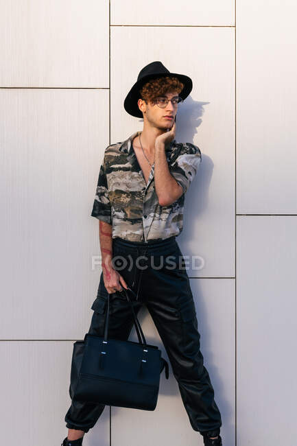 Hombre joven y vanidoso en ropa elegante con bolso de señora de pie en la pared de azulejos mientras mira hacia otro lado - foto de stock