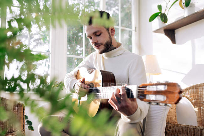 Contemplativo músico masculino com tatuagens tocando guitarra clássica enquanto sentado em poltrona e olhando para a janela em casa — Fotografia de Stock