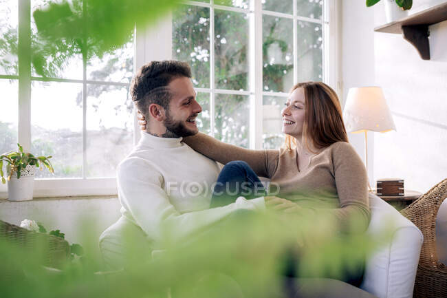 Sonriente joven hembra acariciando el pelo de hombre tatuado amado mientras descansa en casa en un día soleado - foto de stock