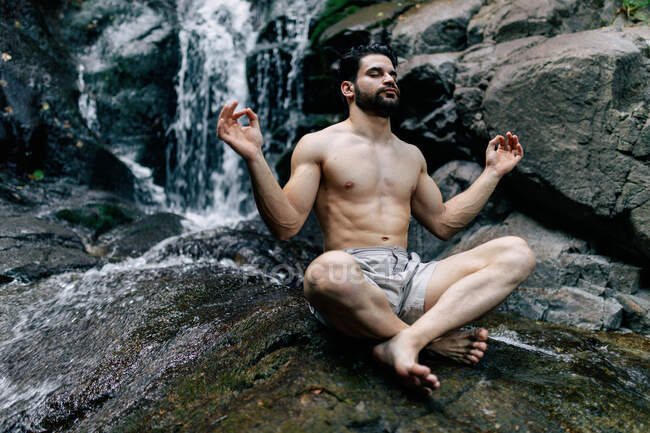 Pacifico hombre sin camisa sentado en Padmasana con las manos de mudra y los ojos cerrados mientras hace yoga y medita sobre roca mojada cerca de la cascada - foto de stock