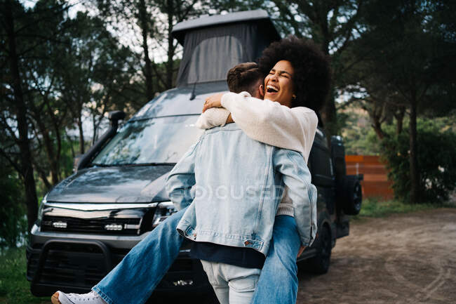 Jovem mulher afro-americana alegre rindo alegremente e abraçando namorado enquanto se divertindo juntos perto de van campista estacionado na floresta verde durante a viagem de verão juntos — Fotografia de Stock