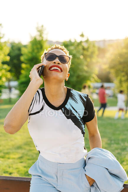 Cheerful African American female with red lips in trendy wear talking on cellphone while looking up in park — Stock Photo
