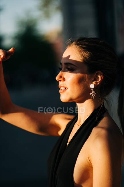Vue latérale de la femme tendance couvrant le visage de la lumière du soleil tout en se tenant dans la rue de la ville — Photo de stock