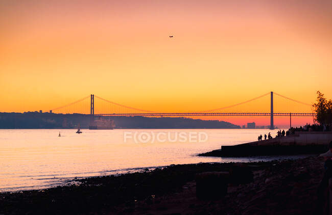 Crowded embankment located near Tagus River with ferry near silhouette of 25 de Abril Bridge against orange sundown sky in Lisbon, Portugal — Stock Photo