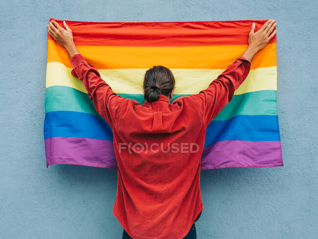Vista trasera de un hombre homosexual anónimo de pie con la bandera del arco iris LGBT cerca de la pared azul en la calle de la ciudad - foto de stock