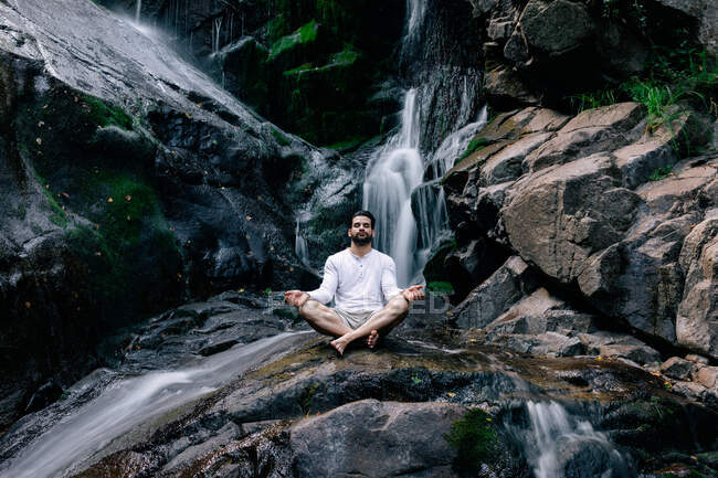 Мирный мужчина сидит в Падмасане с мудрой и закрытыми глазами, занимаясь йогой и медитируя на мокрой скале возле водопада — стоковое фото