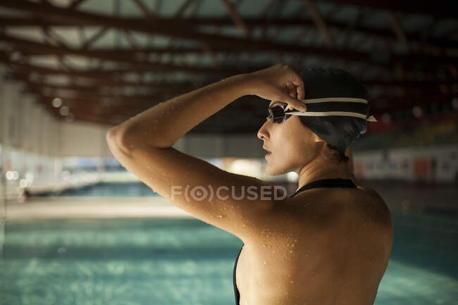 Joven hermosa mujer en la acera de la piscina cubierta, con traje de baño negro, se pone las gafas de baño, vista lateral - foto de stock