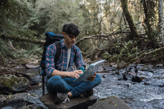 Mochilero masculino viajero con bebida de agua y mapa de papel sentado en la roca cerca del río en el bosque y mirando hacia arriba - foto de stock