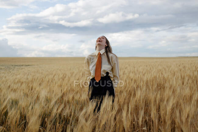 Positive young female in white shirt and red tie standing with hands behind back among wheat spikes in countryside — Stock Photo