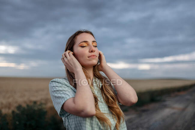 Стильная молодая женщина с закрытыми глазами касается длинных волос на проезжей части под облачным небом в сумерках — стоковое фото