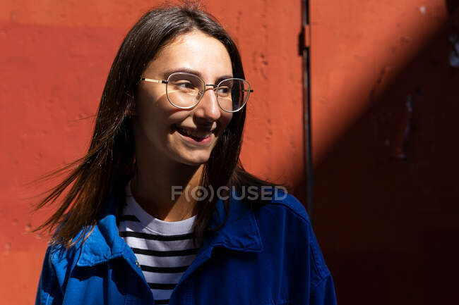 Positive female in stylish outfit looking away on colorful background of wall of building on sunny day in city street — Stock Photo