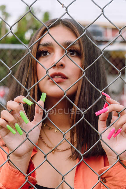 Stylish female with bright long manicure standing near metal fence and looking away with interest — Stock Photo