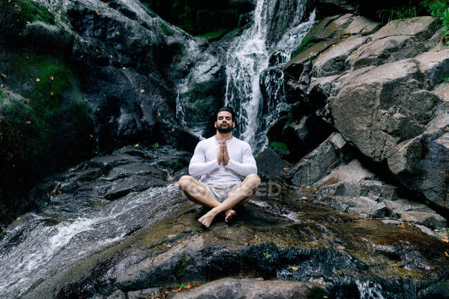 Tranquil male sitting in Lotus pose with prayer hands on rock near waterfall and meditating while doing yoga with closed eyes — Stock Photo