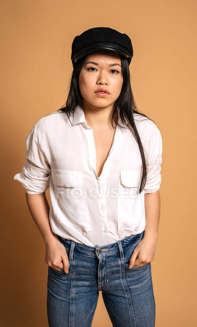 Вид спереди на модную азиатскую модель в белой рубашке и джинсах с руками в джинсовых карманах на бежевом фоне и смотрящую в камеру — стоковое фото