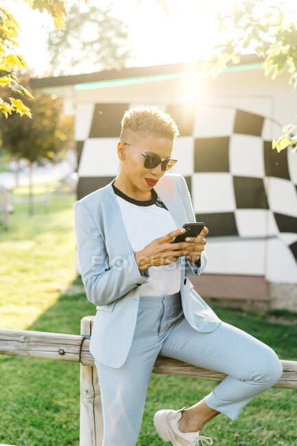 Content mature African American female in modern sunglasses using cellphone in park in back lit — Stock Photo