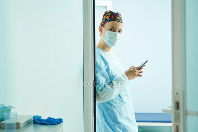Vista lateral del médico femenino en uniforme médico y mensajes de texto de máscara estéril en el teléfono celular en la clínica - foto de stock