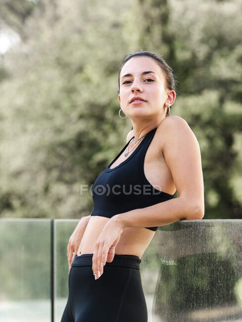 Вид сбоку бегуньи в спортивной одежде, опирающейся на стеклянный забор после тренировки в летний день — стоковое фото