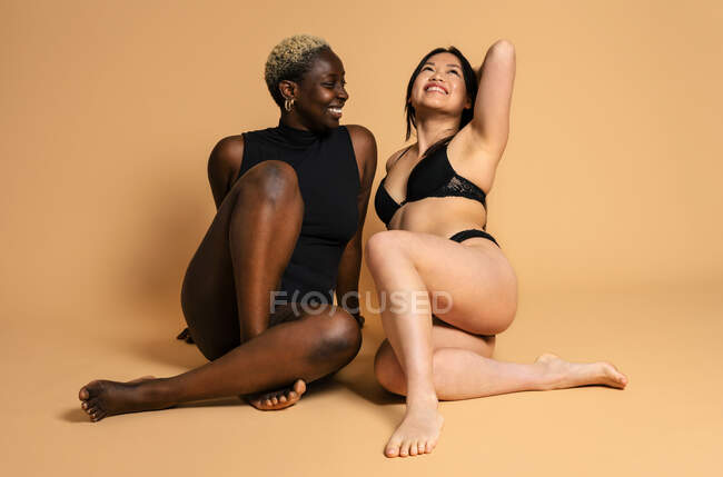 Glad femmes multiraciales portant de la lingerie noire assis sur fond beige en studio et souriant pour le concept de positivité du corps — Photo de stock