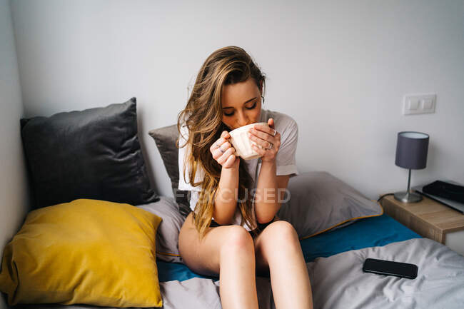 Нежная женщина в трусиках и футболке сидит на мягкой кровати и пьет горячий напиток из чашки во время охлаждения дома — стоковое фото