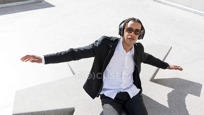 High angle of young ethnic male with braided hair dressed in stylish suit and sunglasses enjoying music through wireless headphones while resting on sunny urban square — Stock Photo