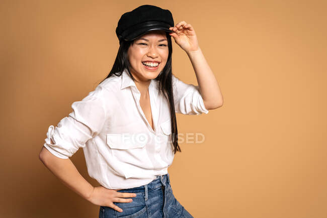 Передній вид моди азіатська щаслива жіноча модель в білій сорочці і джинсах з руками в кишенях на бежевому тлі і дивлячись на камеру — стокове фото
