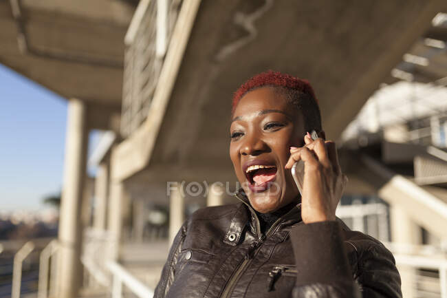 Vista lateral de una hermosa mujer afro negra hablando con su teléfono inteligente mientras sonríe con un fondo borroso en un día soleado - foto de stock