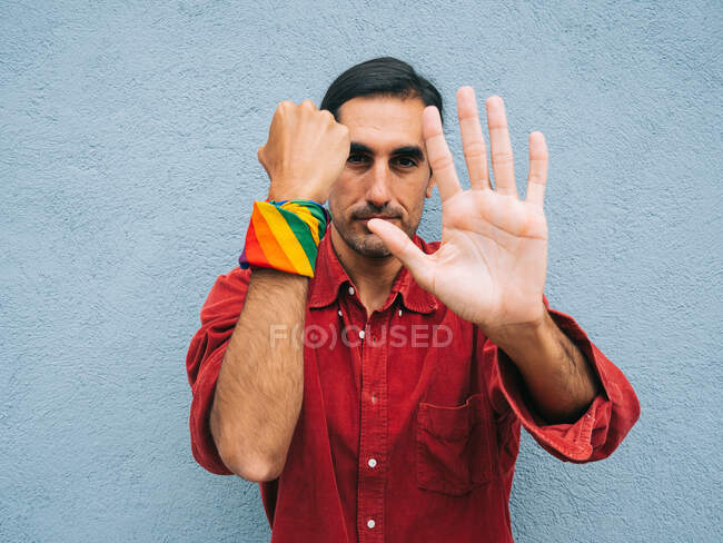 Etnico gay maschio con arcobaleno bandana su mano mostrando stop segno su grigio sfondo in strada e guardando fotocamera — Foto stock