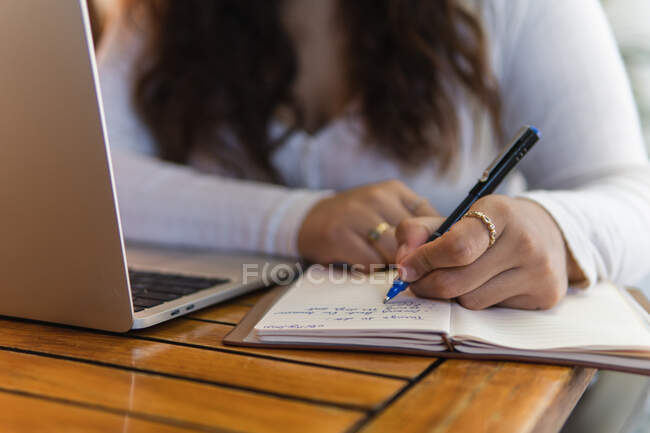 Обрезанная неузнаваемая студентка делает заметки в планировщике во время работы над ноутбуком, готовясь к университетскому экзамену в уютном кафе — стоковое фото