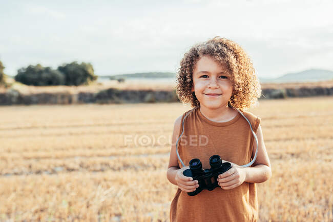 Delighted ethnic child with Afro hairstyle looking at camera holding binoculars standing in dried filed in summer on sunny day and having fun — Stock Photo