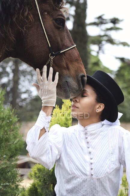 Serious adult black female in hat and elegant outfit with gloves petting brown horse with closed eyes near green plants and trees in countryside — Stock Photo
