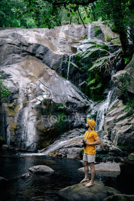 Мужчина исследователь в желтой одежде и с винтажной фотокамерой, стоящей на заднем плане водопада в лесу и смотрящей в сторону — стоковое фото