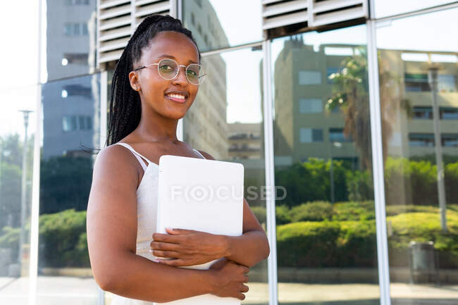 Delighted African American female freelancer with braids and white netbook standing in street in Barcelona and looking at camera — Stock Photo