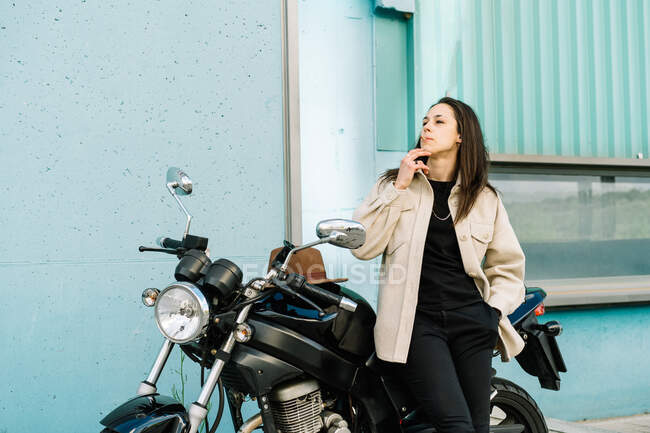 Femme confiante motard penché sur la moto garée sur le bord de la route en ville et fumer la cigarette tout en regardant loin — Photo de stock
