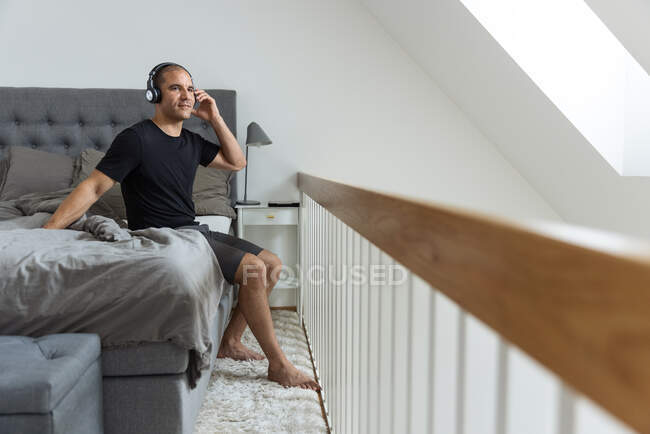 Vista lateral do homem tranquilo colocando fones de ouvido e ouvindo música enquanto está sentado na cama depois de acordar no quarto e desfrutar da manhã — Fotografia de Stock