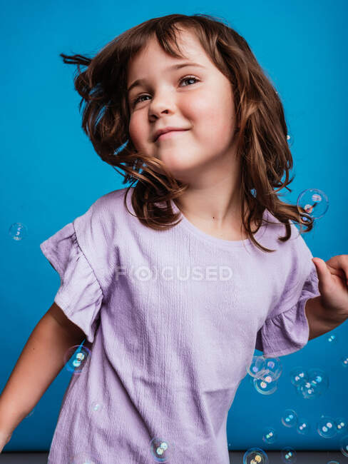Criança despreocupada no vestido dançando e sorrindo em estúdio com bolhas de sabão flutuantes em fundo azul vívido — Fotografia de Stock