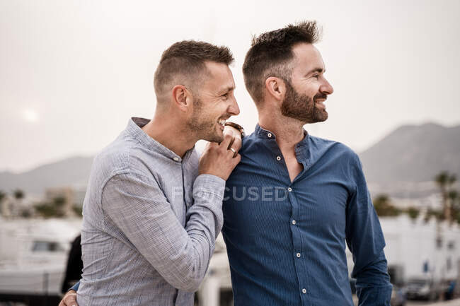 Couple of homosexual men in shirts embracing while looking away on harbor against ocean and mountain — Stock Photo