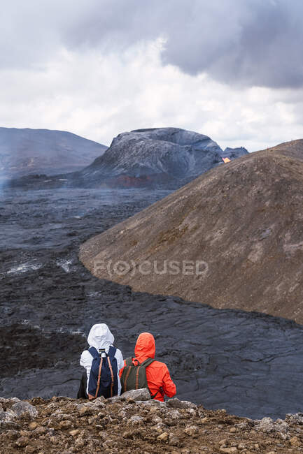 Vista posteriore di viaggiatori anonimi in capispalla contemplando lava di Fagradalsfjall dal monte sotto il cielo nuvoloso in Islanda — Foto stock