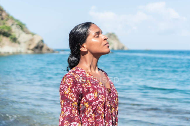 Ethnic female tourist in sundress standing with closed eyes on sandy coast against ocean and mounts in sunlight — Stock Photo
