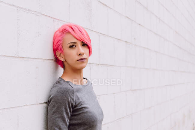Jeune femme pensive avec des cheveux teints en vêtements décontractés regardant la caméra tout en se tenant près du mur blanc — Photo de stock