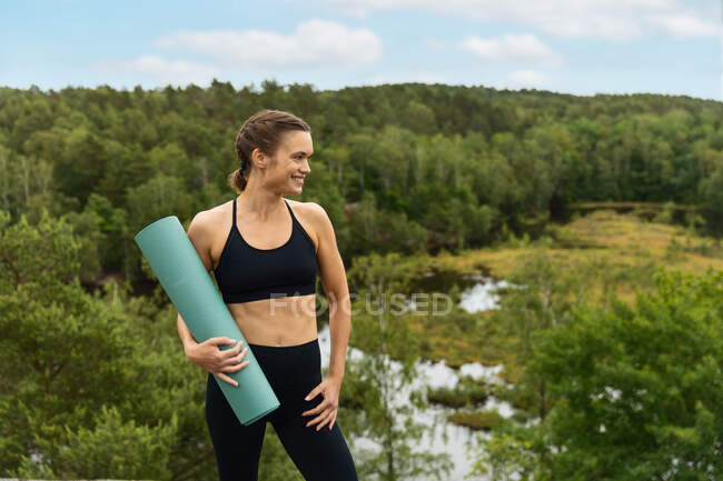 Heureuse jeune femme en tenue de sport noire portant roulé tapis et détournant les yeux avec sourire avant la séance de yoga dans une campagne luxuriante — Photo de stock