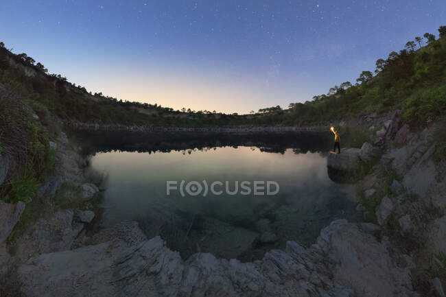 Anónimo viajero con antorcha contemplando laguna entre montañas bajo cielo estrellado al atardecer en España - foto de stock