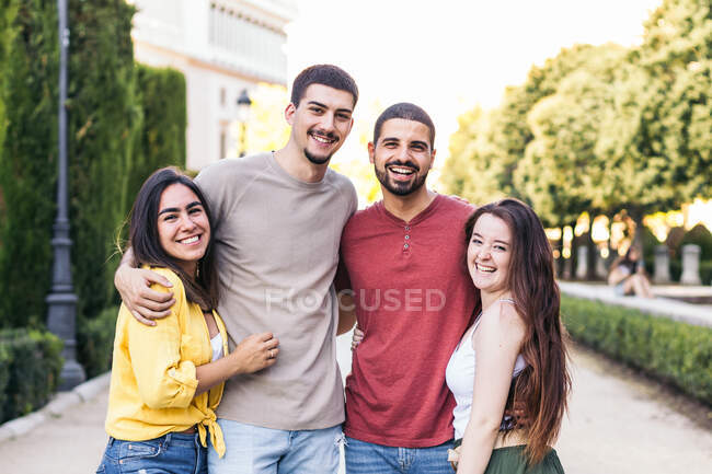 Company of cheerful friends embracing and standing along street together in park during weekend — Stock Photo