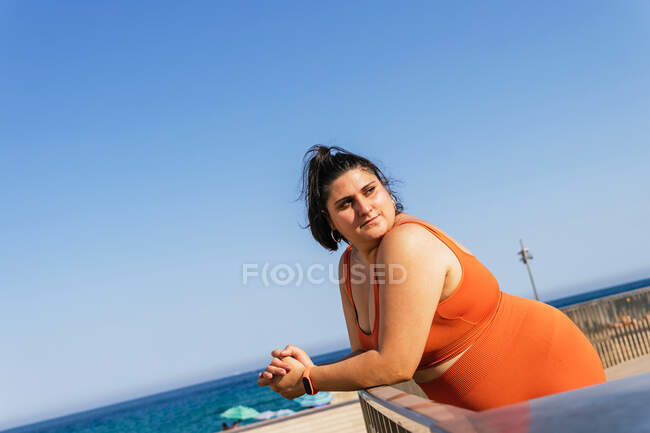 Вид збоку на надмірну вагу етнічної жінки-спортсменки з загорнутими руками, дивлячись подалі, спираючись на паркан на океан у місті — стокове фото